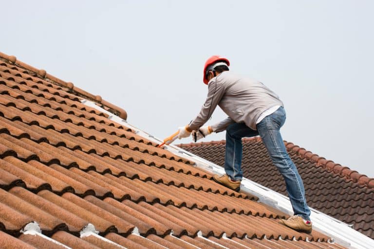 Roofer wearing a safety helmet and gloves, working on a clay tile roof near the ridge with a roofing tool.